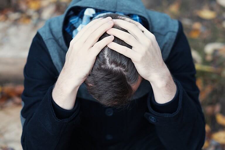 Young man covers his face with his hands in grief or pain. He is sitting outdoors