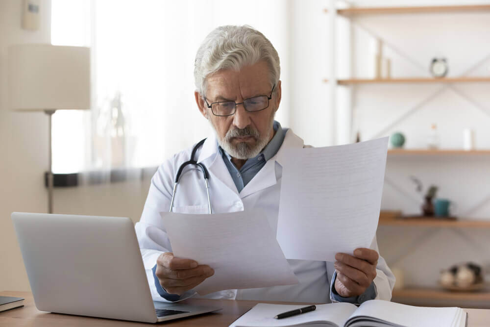 Serious professional senior elderly doctor doing paperwork checking medical documents at workplace.