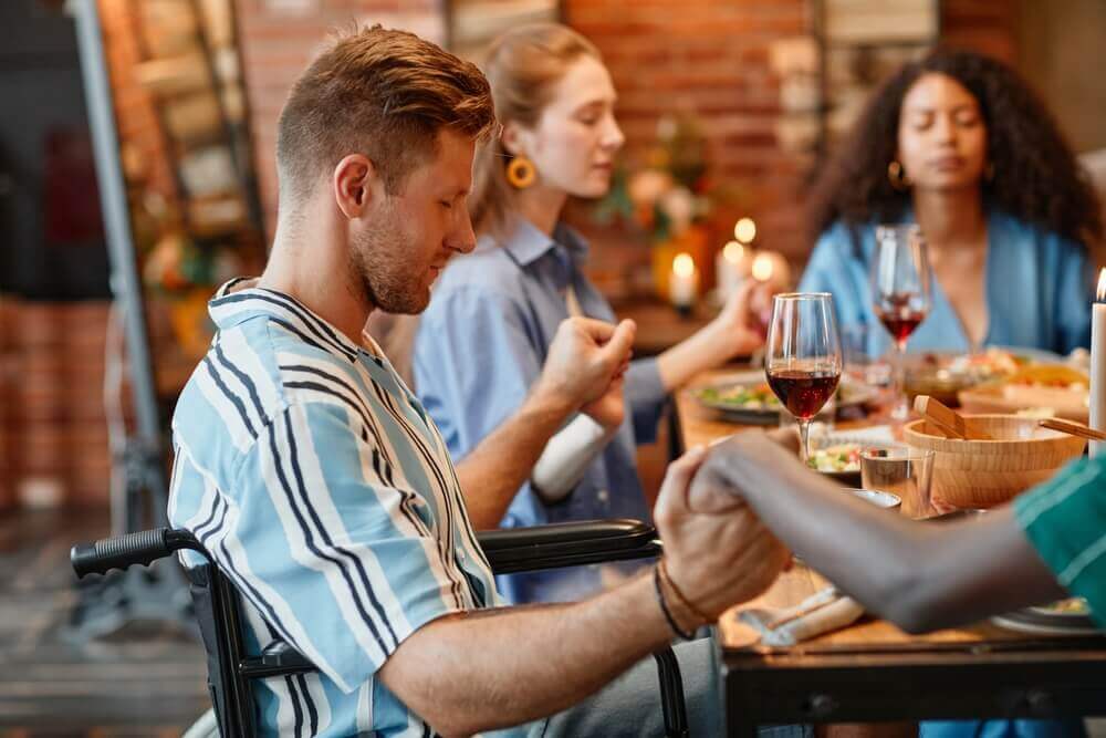 Side view portrait of young man in wheelchair holding hands with friends and saying grace at dinner party in cozy setting sitting at the table and celebrating holiday.