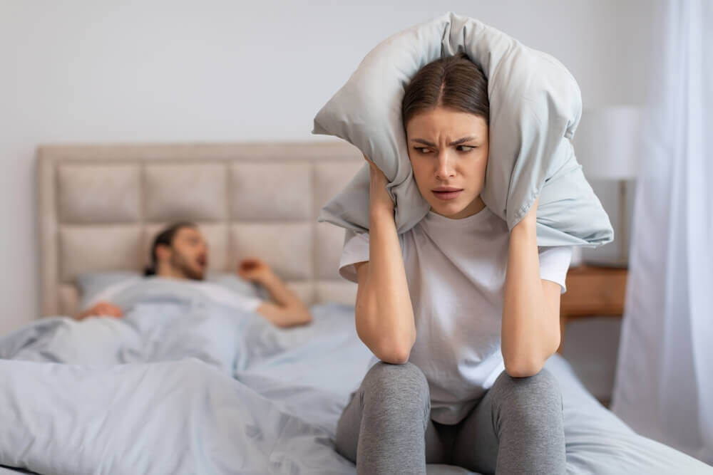 young woman covers her ears with pillow to block out the loud snoring of man sleeping in the background.