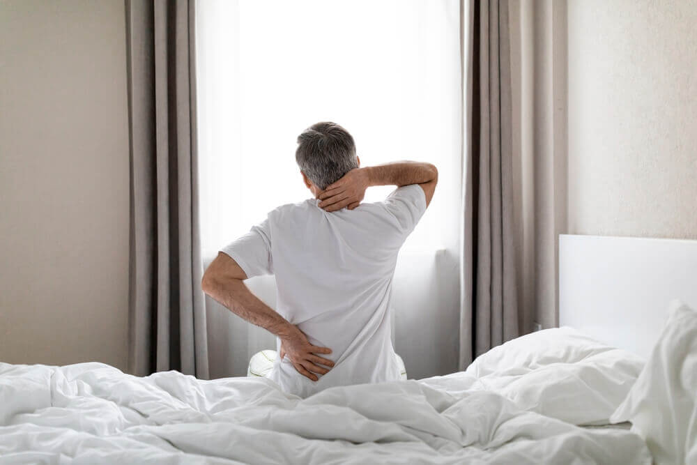 grey-haired man wearing white pajamas sittting on bed at home in the morning, touching lower back and neck, suffering from muscle strain.