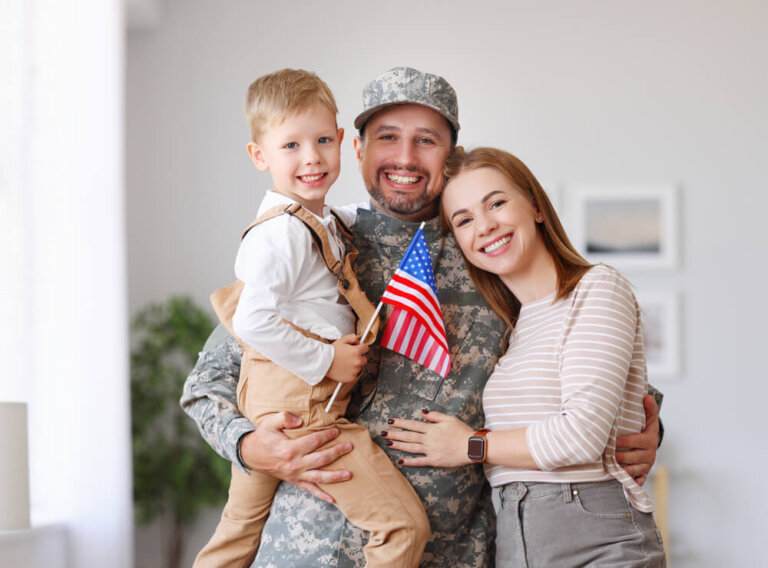 Happy young man father in military uniform reunited with his beautiful american family, hugging with smiling wife and cute kid son with flag of United
